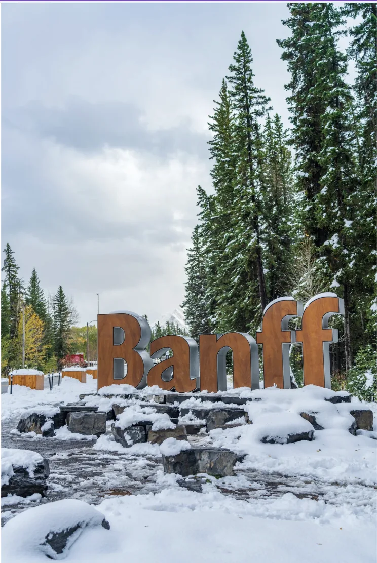 Snow-covered Banff welcome sign surrounded by evergreen trees.